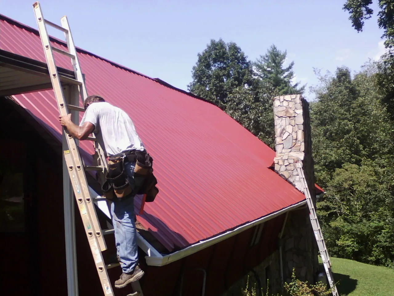 Roofer on a ladder beside a red metal roof and stone chimney — Soffit & Fascia Repair by Roofing Brothers in New Ipswich